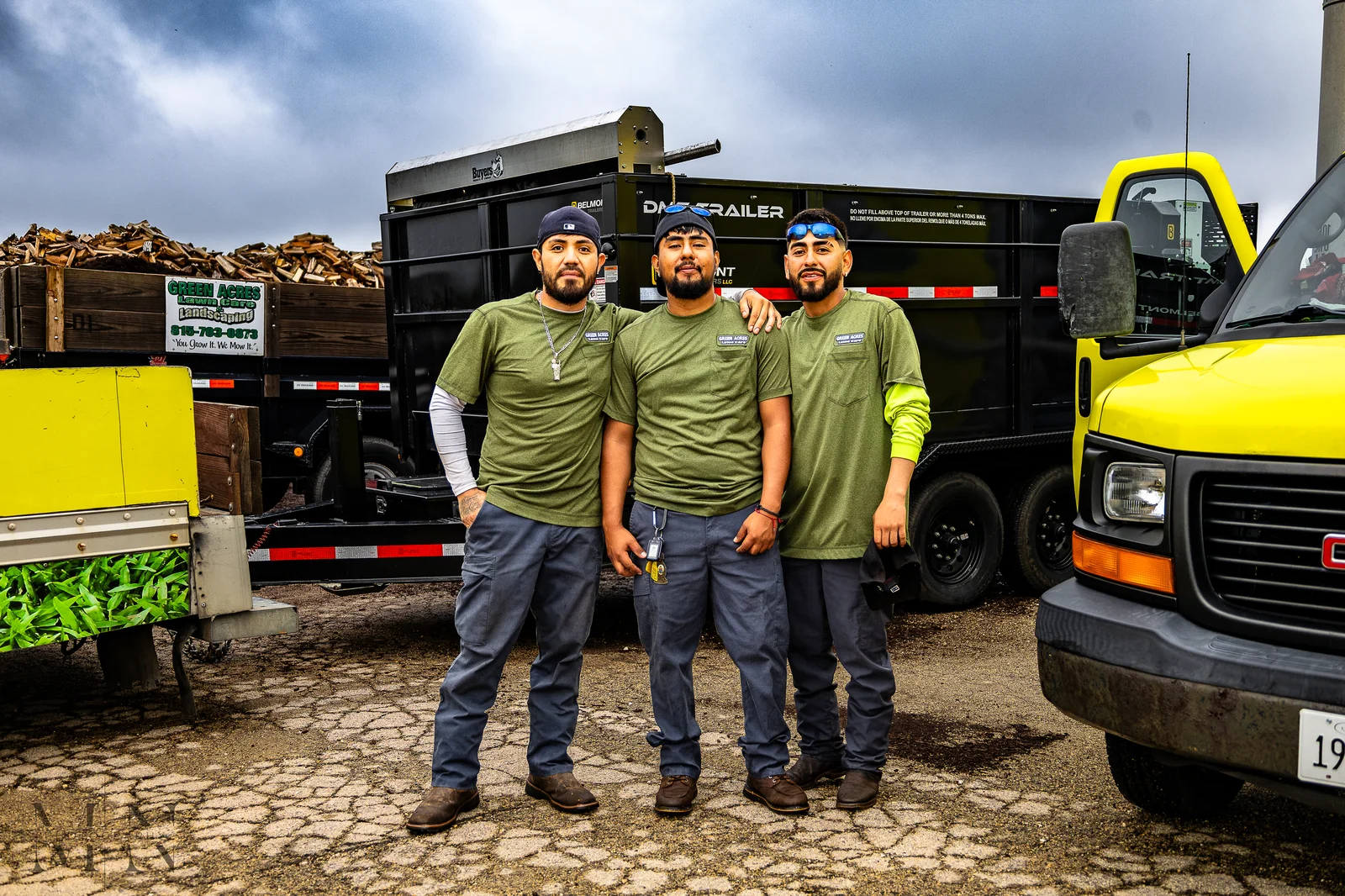 Green Acres landscaping crew members standing in front of company dump trailer and yellow truck at work site
