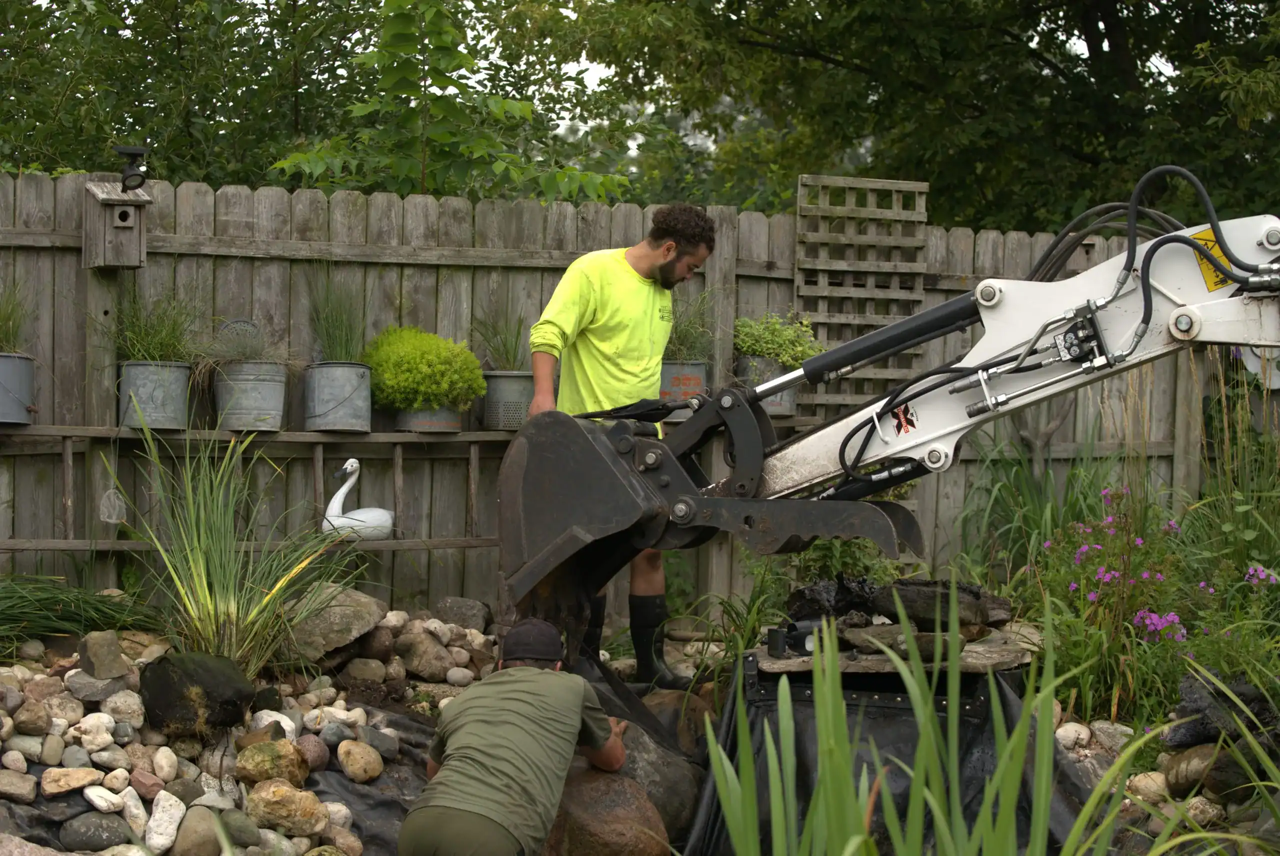 Green Acres crew member in safety yellow shirt operating mini excavator for pond maintenance and landscaping service